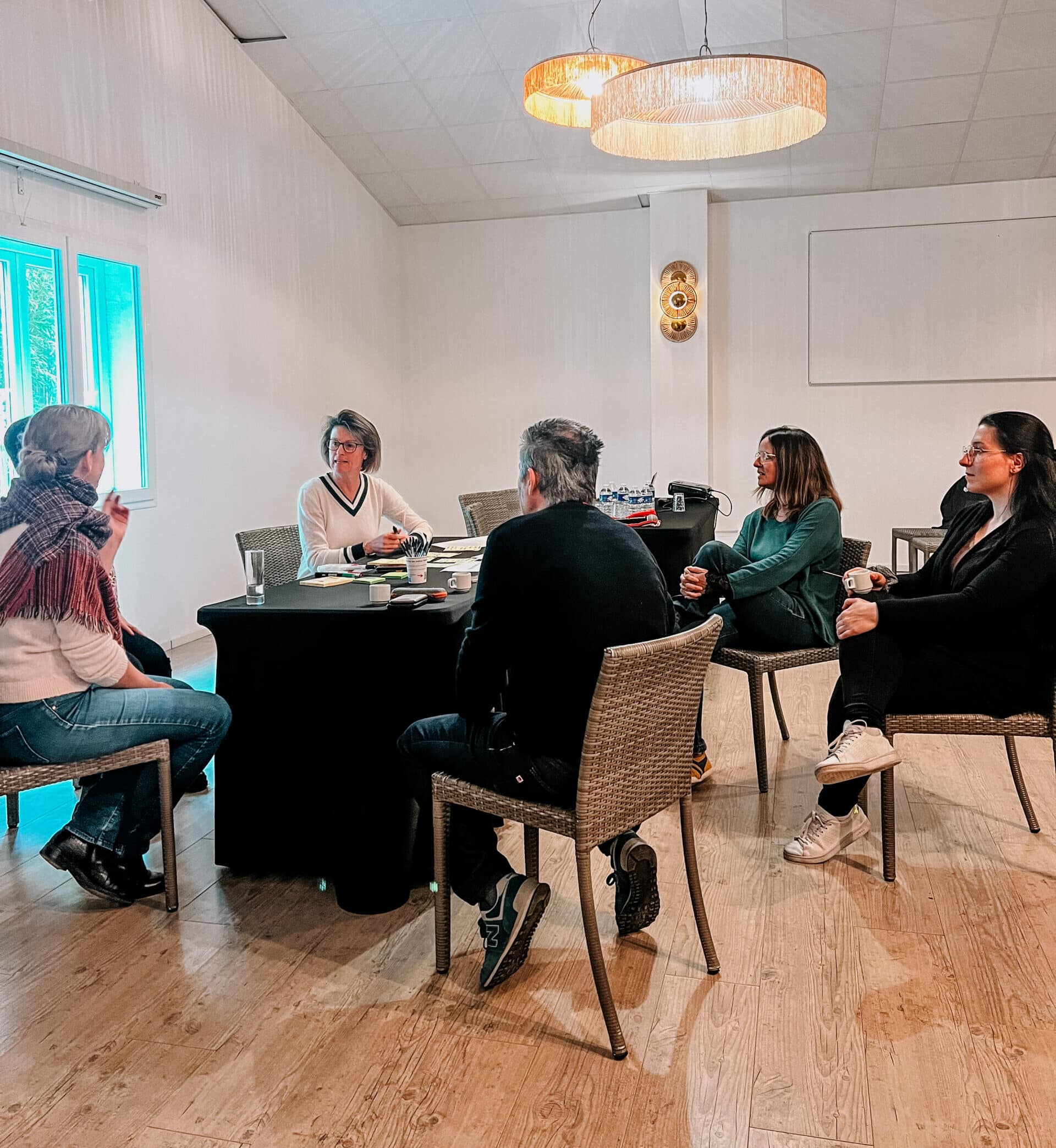 Discussion d'équipe professionnelle dans une salle de réunion lumineuse. Cinq participants assis sur des chaises en rotin autour d'une table.
