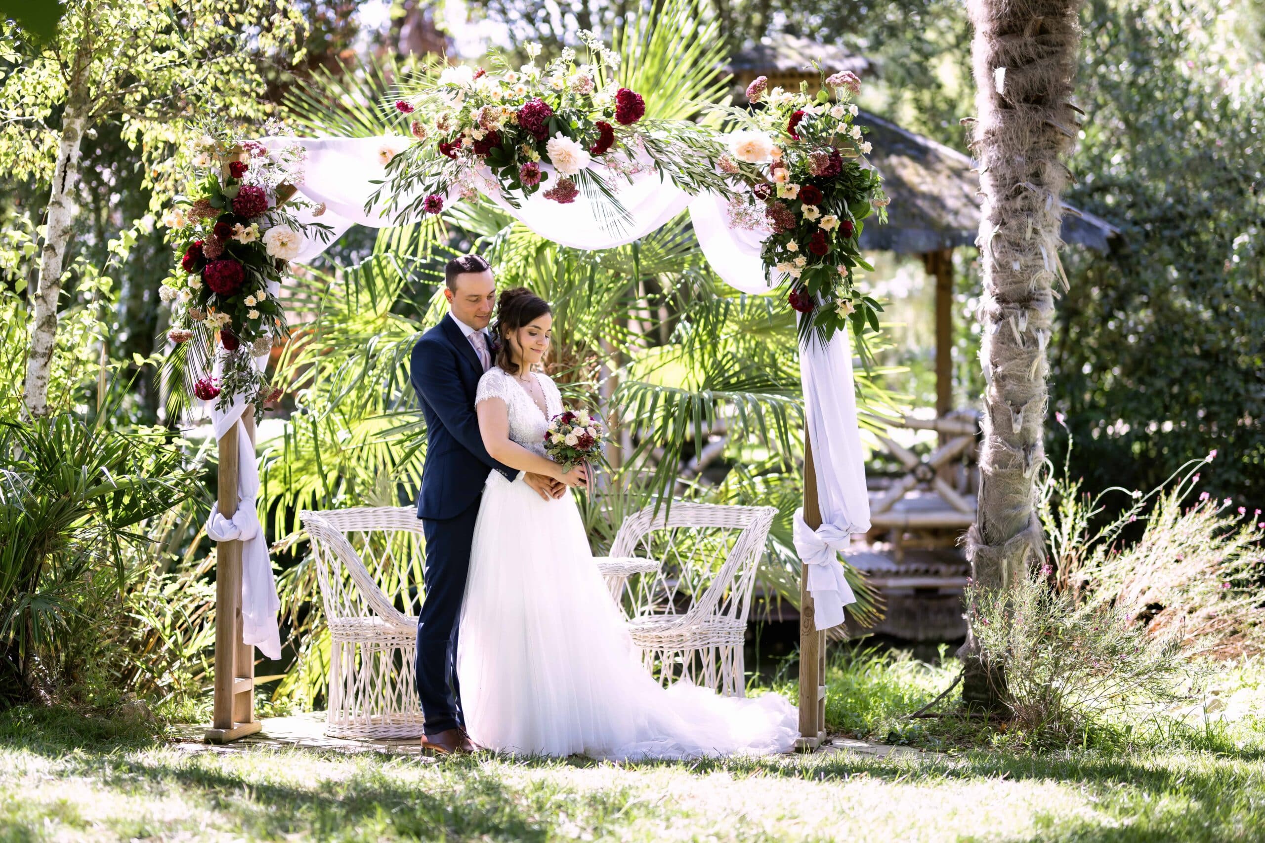Élégant couple de mariés dans un jardin luxuriant, sous une arche décorée de fleurs bordeaux et crème. Moment intime.
