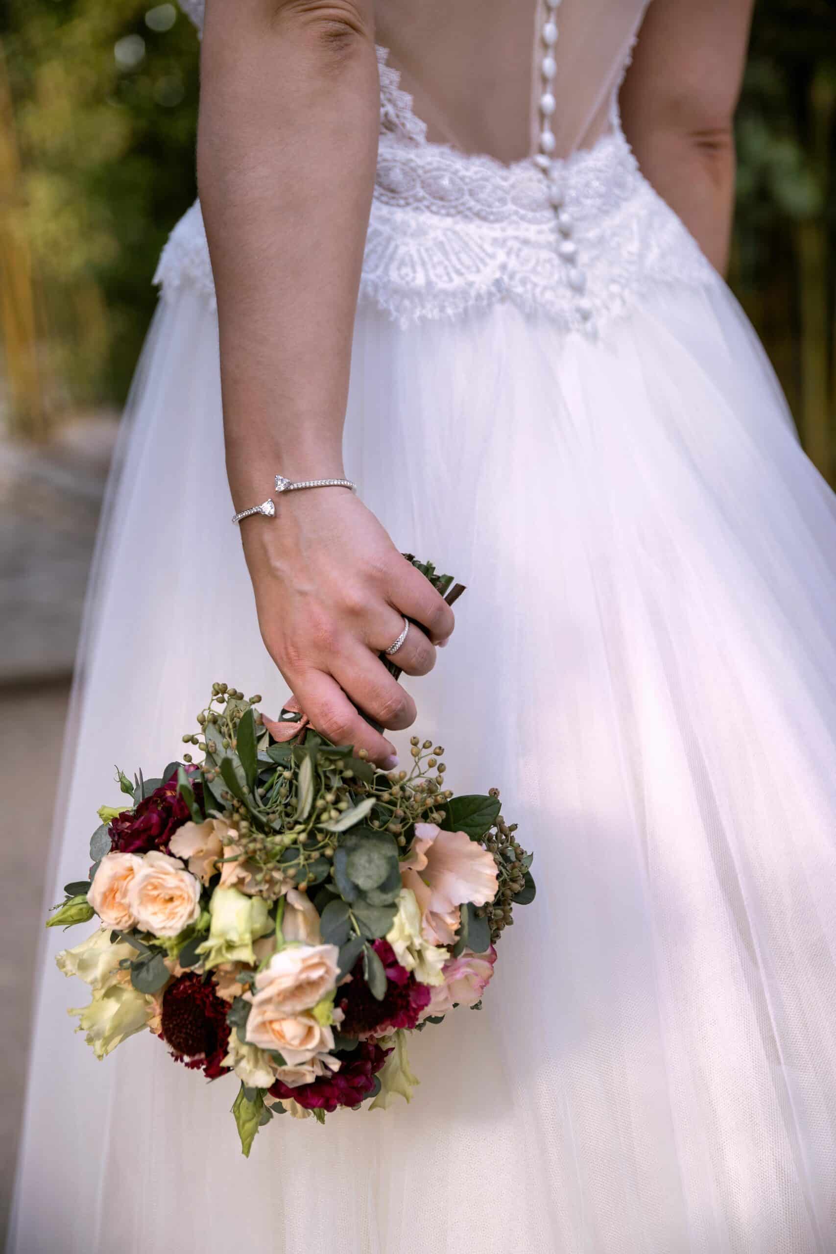 Mariée en robe de tulle dos nu, dentelle et boutons perlés, tenant un bouquet de roses pêche et bordeaux, bracelet diamant.