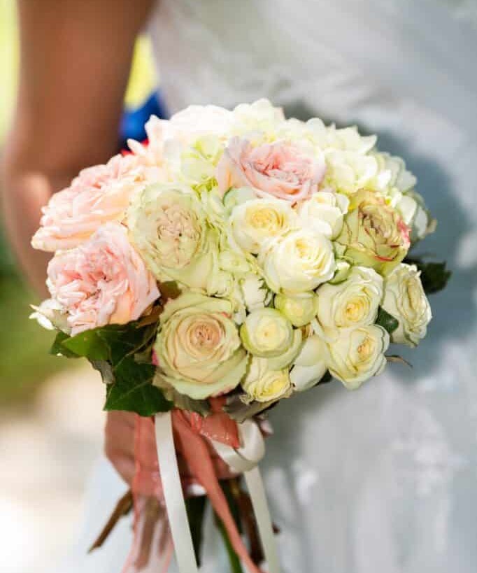 Magnifique bouquet de mariée composé de roses blanc cassé et rose pâle, tenu par la mariée et orné de longs rubans pêche.