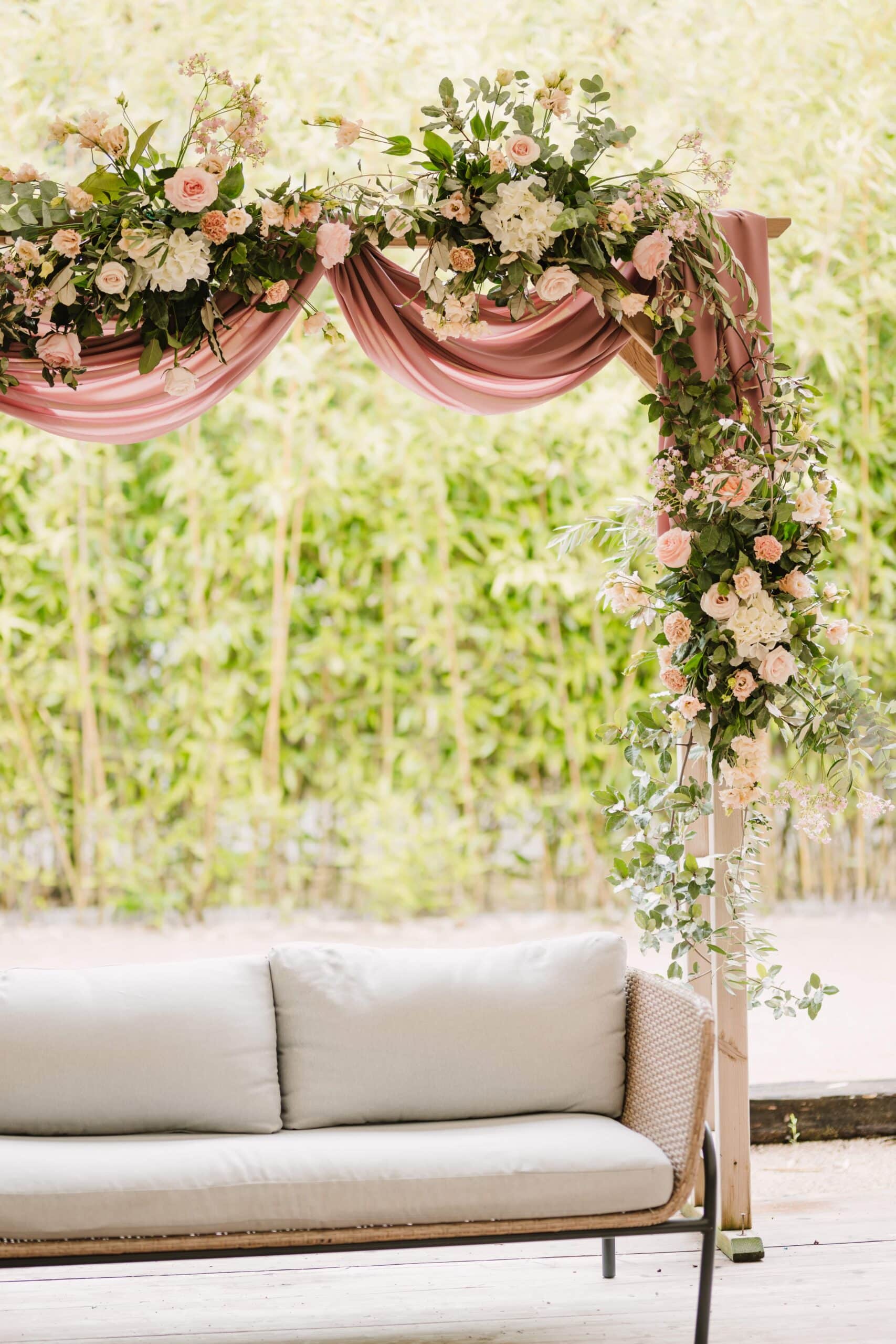 Canapé extérieur élégant sous une arche de fleurs roses et blanches avec drapé romantique en plein air.
