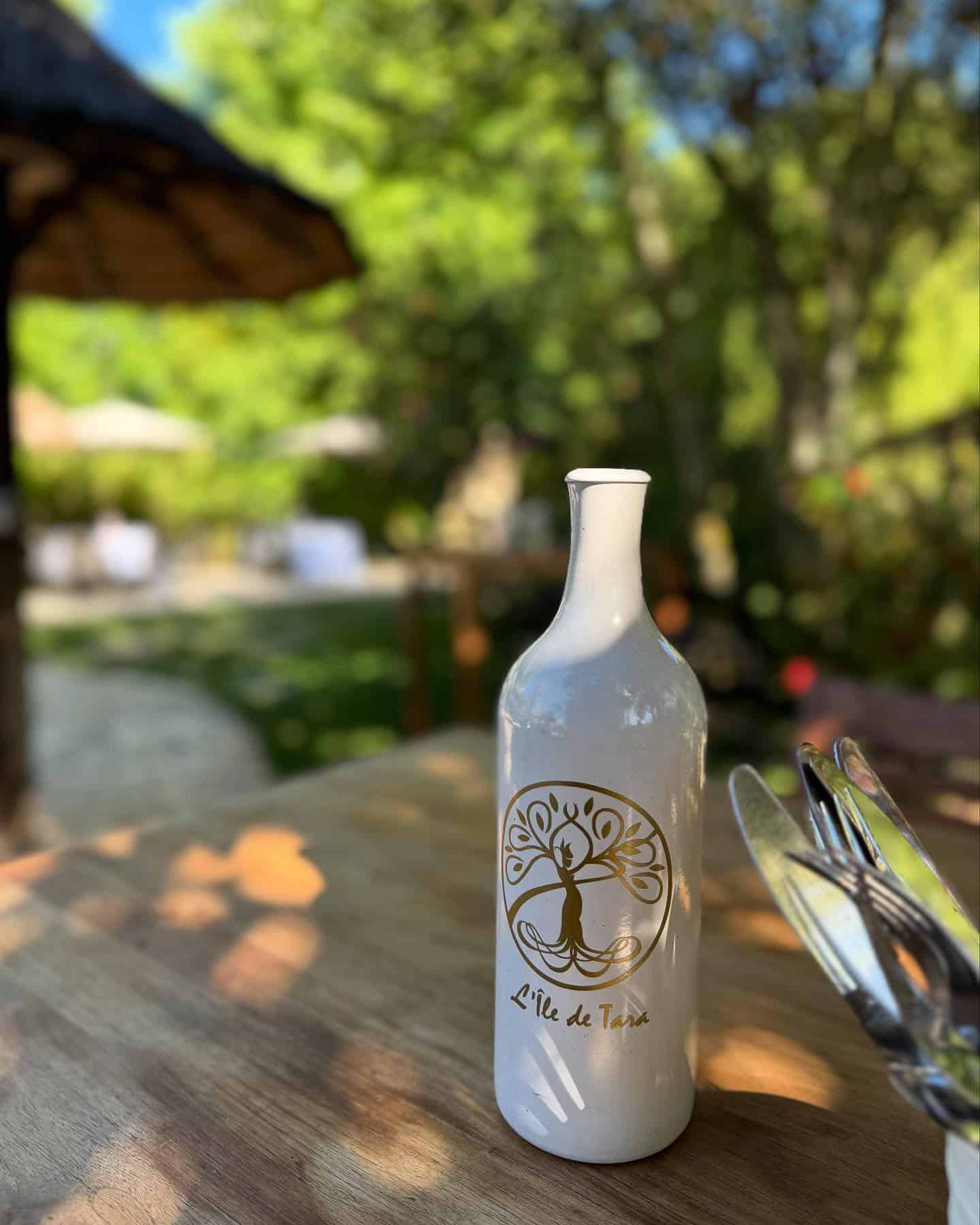 Bouteille en céramique blanche L'île de Tara avec logo doré sur une table en bois en terrasse ensoleillée.