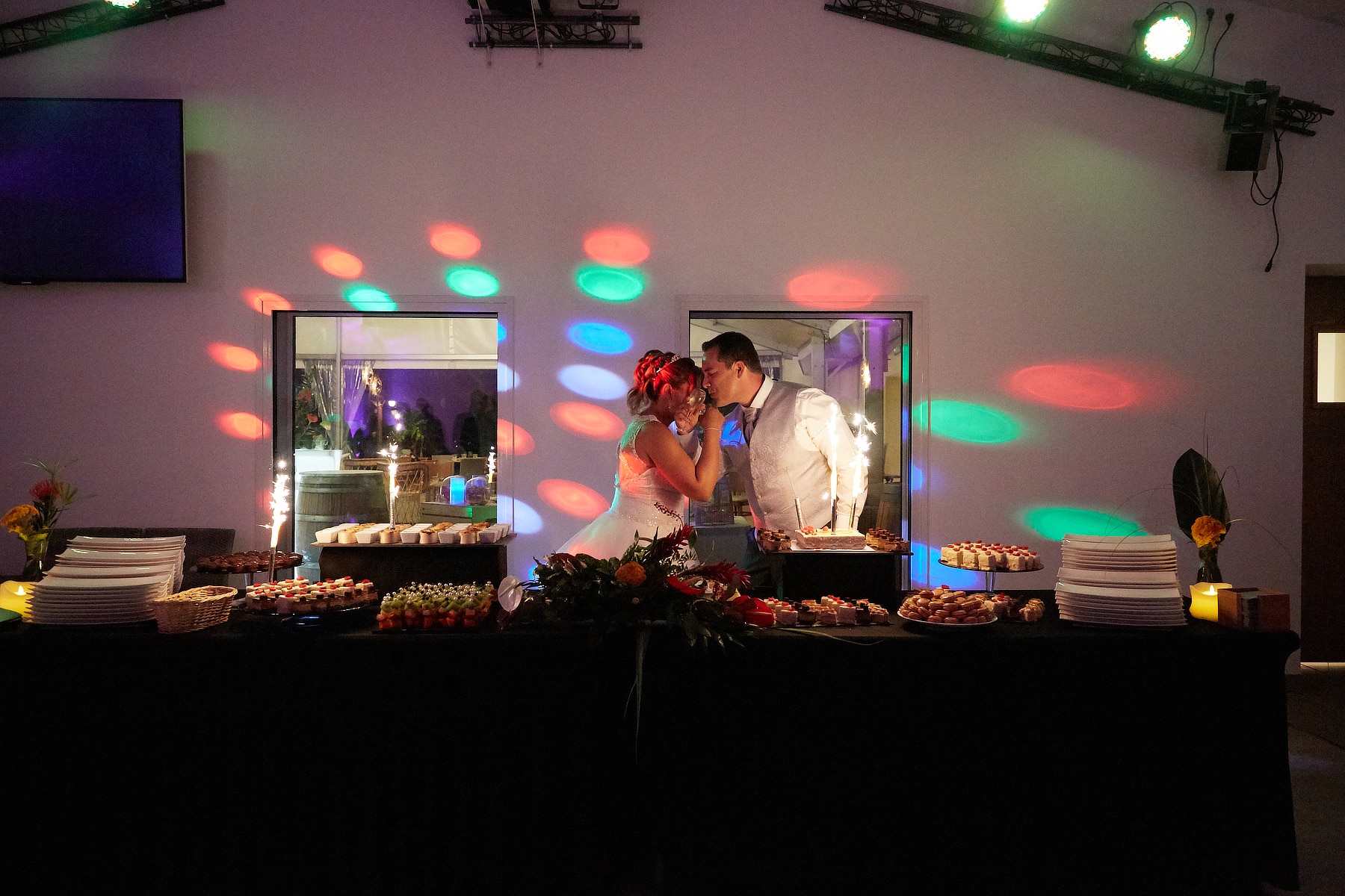 Mariés devant un buffet de desserts festif avec feux de Bengale et jeux de lumières colorés lors d'une réception.