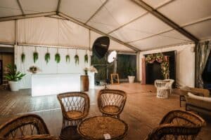 Salon événementiel sous chapiteau avec bar lumineux, chaises en osier et espace photobooth avec décor fleuri Lencarta.