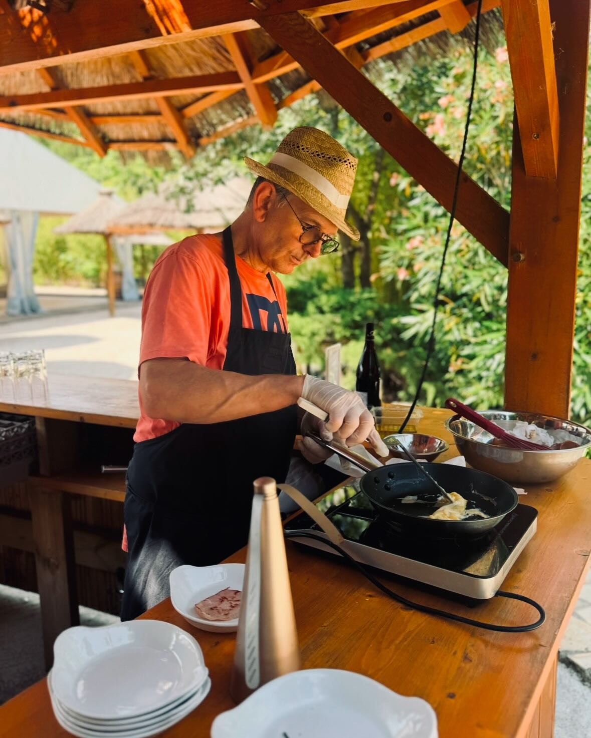 Chef avec chapeau de paille cuisinant en plein air sous un pavillon en bois sur une plaque à induction.