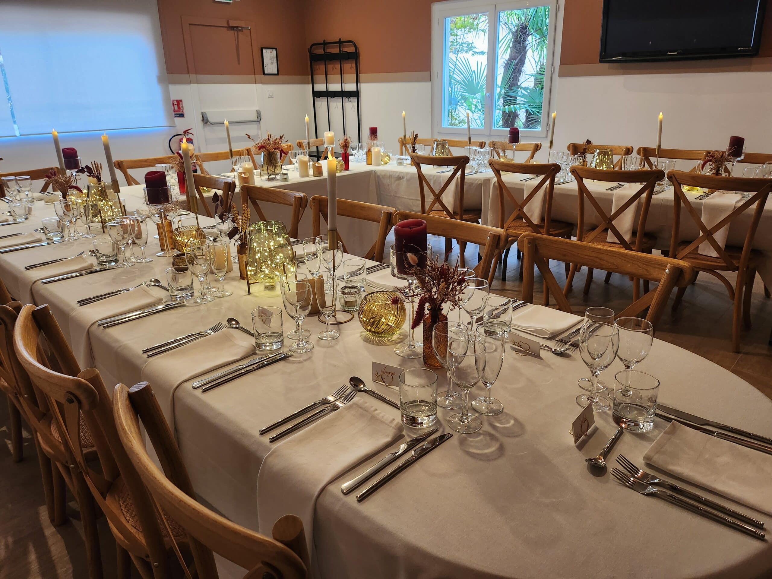 Table de banquet avec bougies bordeaux, guirlandes lumineuses et chaises en bois pour une réception de mariage élégante.