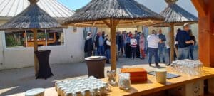 Groupe discutant sous des parasols en chaume lors d'un événement extérieur avec buffet café au premier plan.