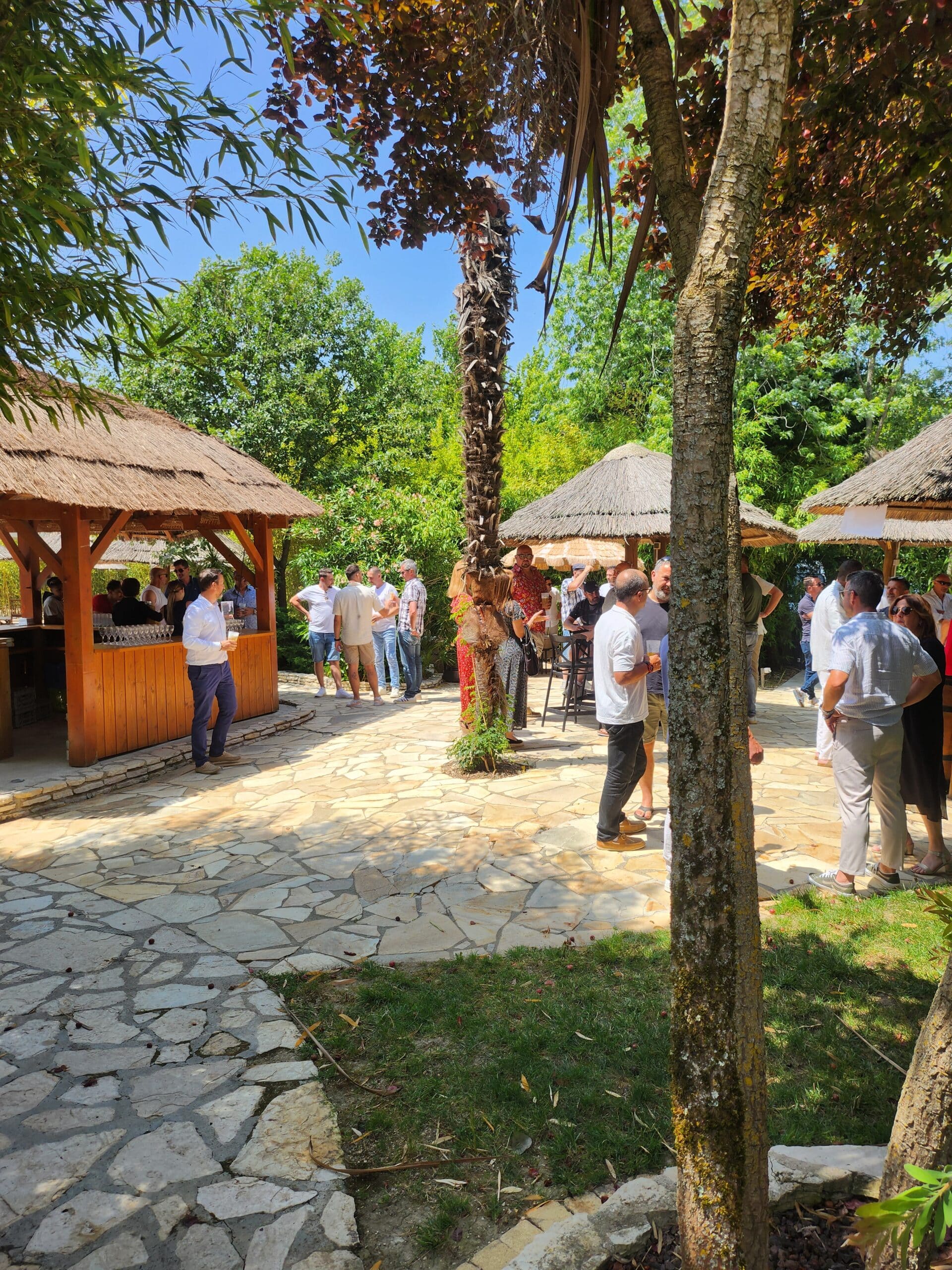 Invités discutant près d'un bar tiki et parasols en paille sur une terrasse en pierre ensoleillée.