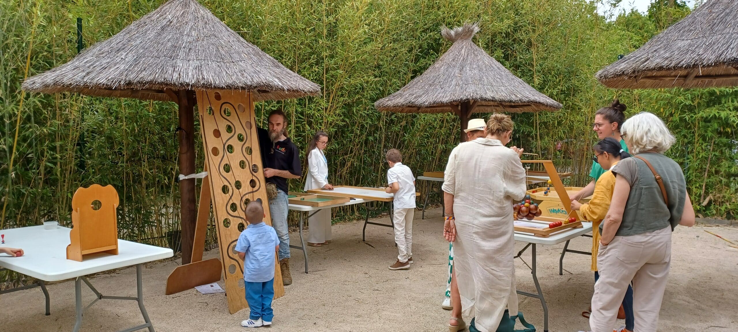 Enfants et adultes s'amusent avec des jeux géants en bois dans un parc arboré de bambous.