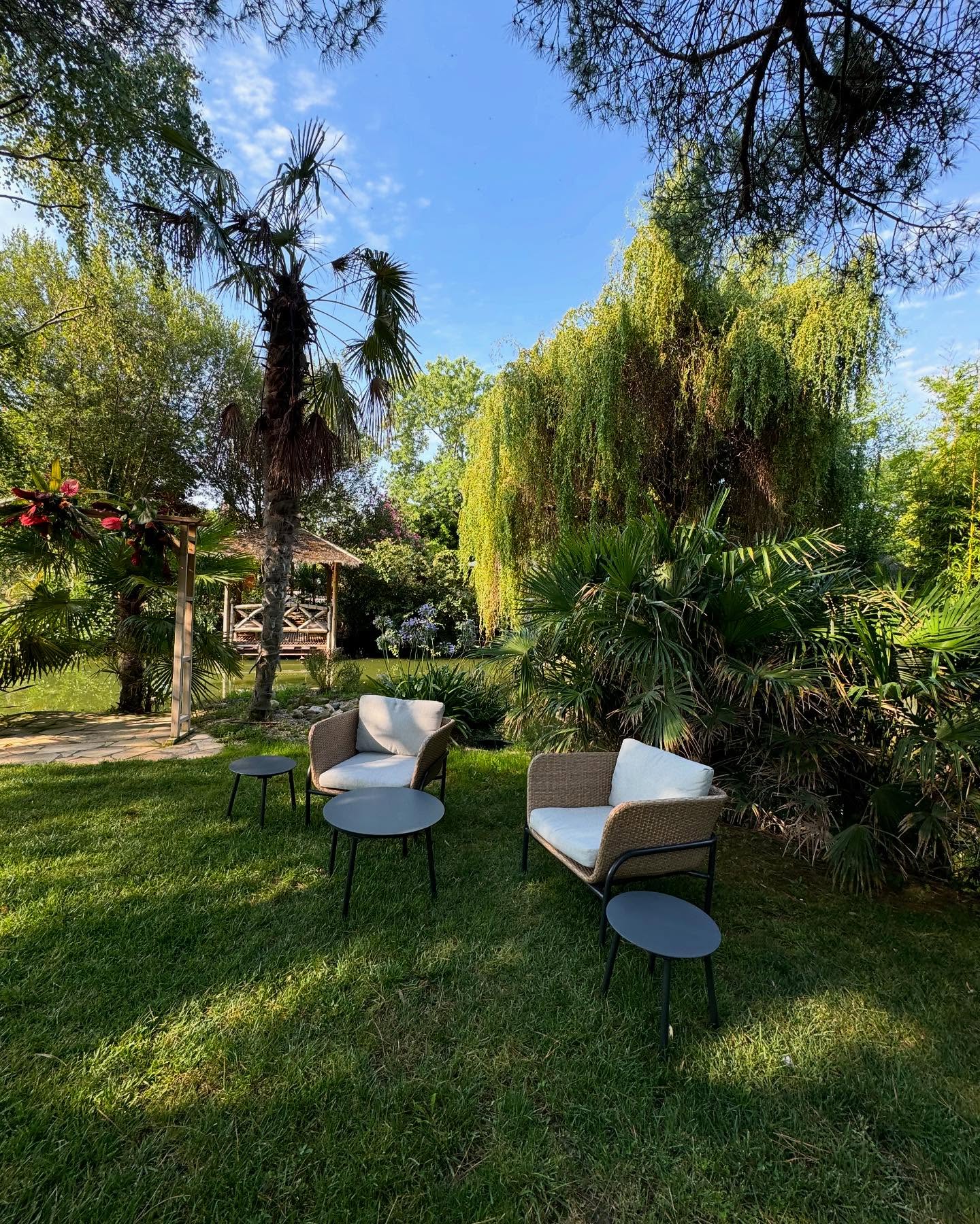 Fauteuils de jardin en rotin sur pelouse avec palmiers et gazebo au bord de l'eau dans un jardin tropical.