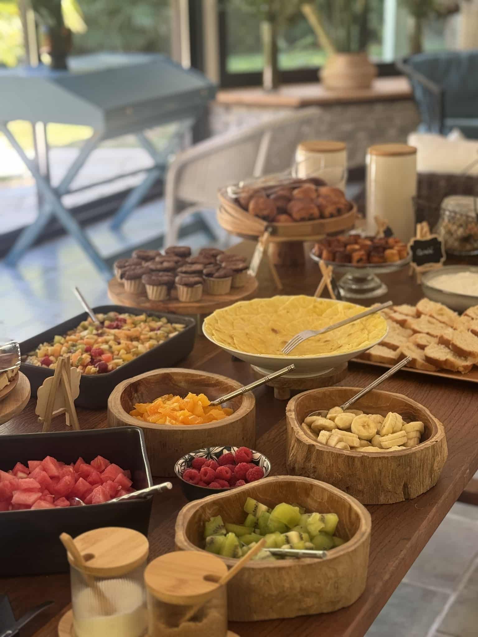 Buffet de petit-déjeuner frais avec fruits variés, viennoiseries et muffins sur une table en bois lumineuse.
