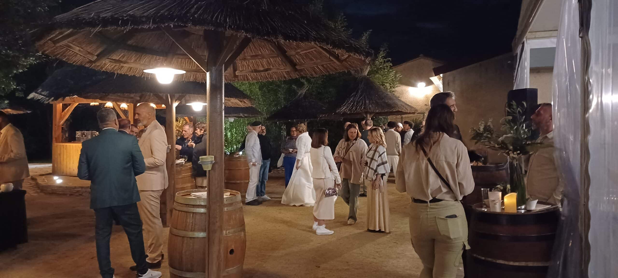 Invités discutant lors d'un cocktail nocturne sous des paillotes exotiques avec tonneaux de vin.