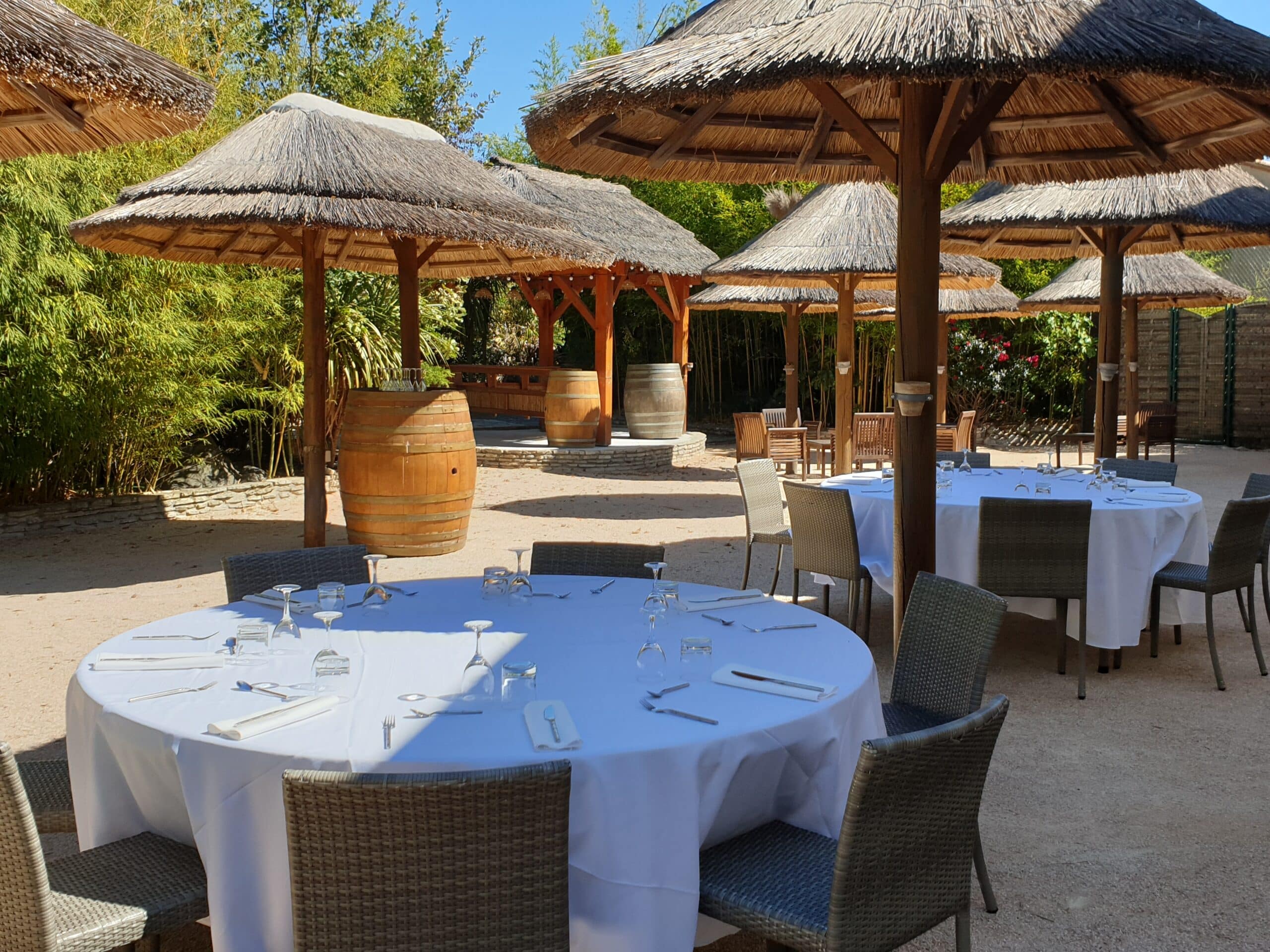 Tables rondes dressées sous des parasols en paille dans un jardin luxuriant avec tonneaux décoratifs.