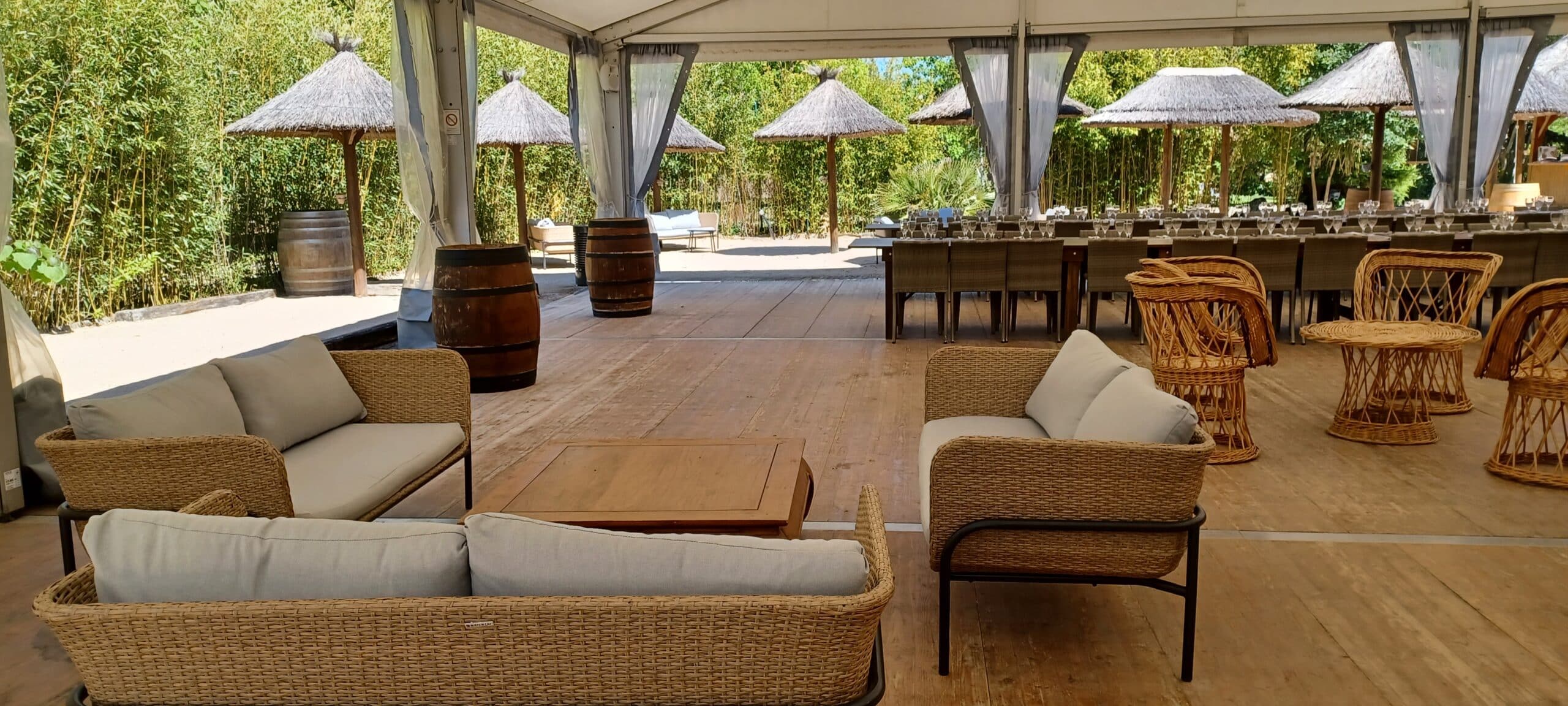 Salon de jardin en osier avec canapés gris, tonneaux décoratifs et paillotes sous une tente de réception.